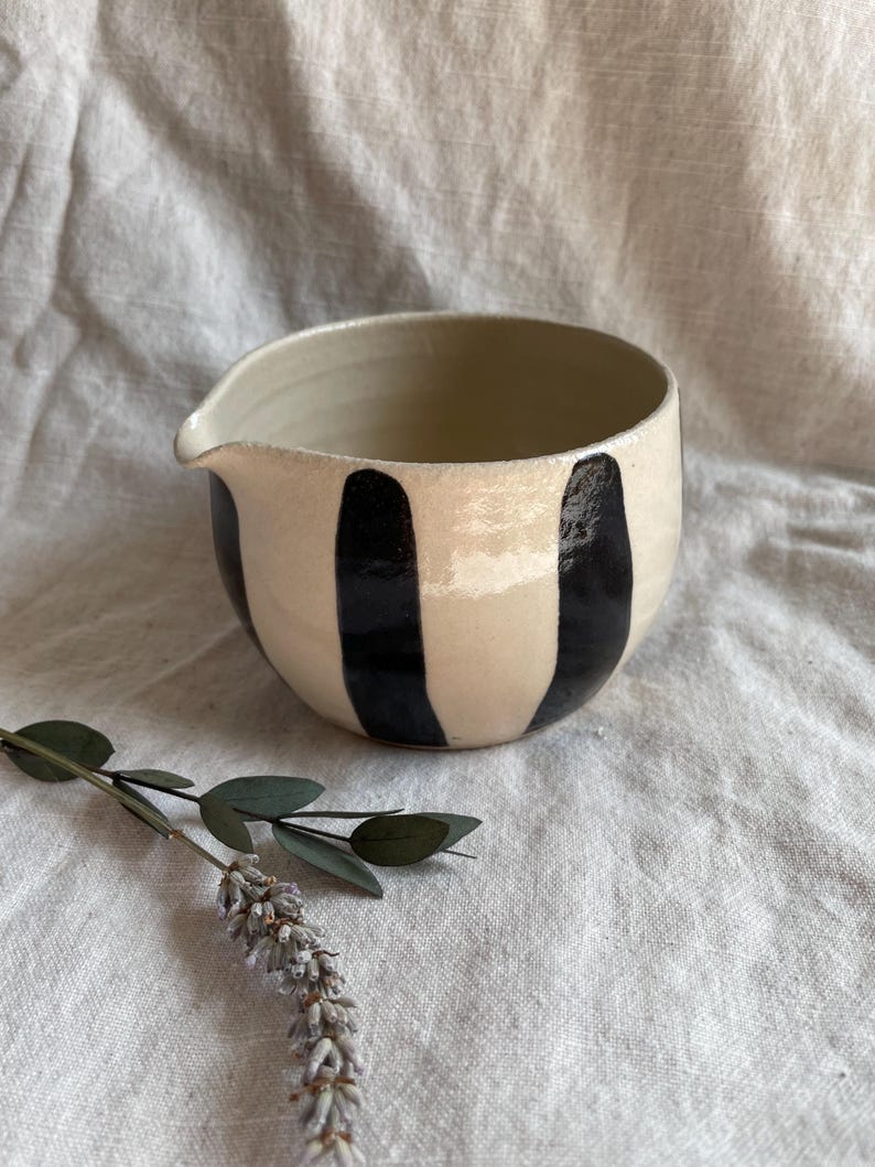 May include: A white ceramic pitcher with black vertical stripes. The pitcher has a rounded shape and a small spout.