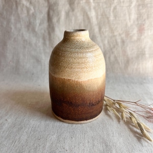 May include: A small, hand-thrown ceramic vase with a brown and cream glaze. The vase has a rounded body and a narrow opening.