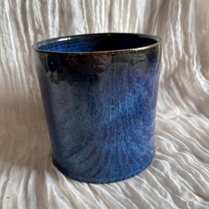May include: A handmade ceramic tumbler with a glossy, deep blue glaze. The cylindrical cup has a dark rim and a textured surface, showcasing a gradient of blue hues. The background is a white, textured fabric.