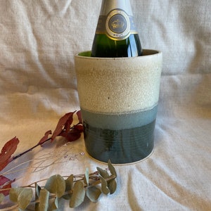 May include: A ceramic wine chiller with a champagne bottle inside. The chiller has a layered design with beige, blue, and dark green bands. Dried foliage accents the scene, set against a neutral fabric backdrop.