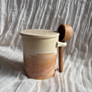 May include: A ceramic jar with a lid and a wooden scoop. The jar is beige at the top, fading to reddish-brown at the base. A wooden scoop is attached to the side. The jar rests on a white, textured surface.