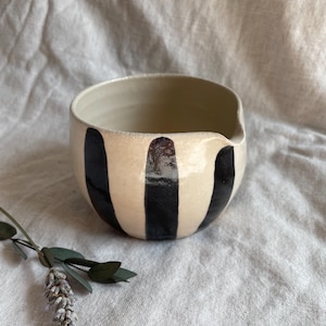 May include: A white ceramic bowl with black vertical stripes. The bowl has a slightly rounded shape and a wide rim.