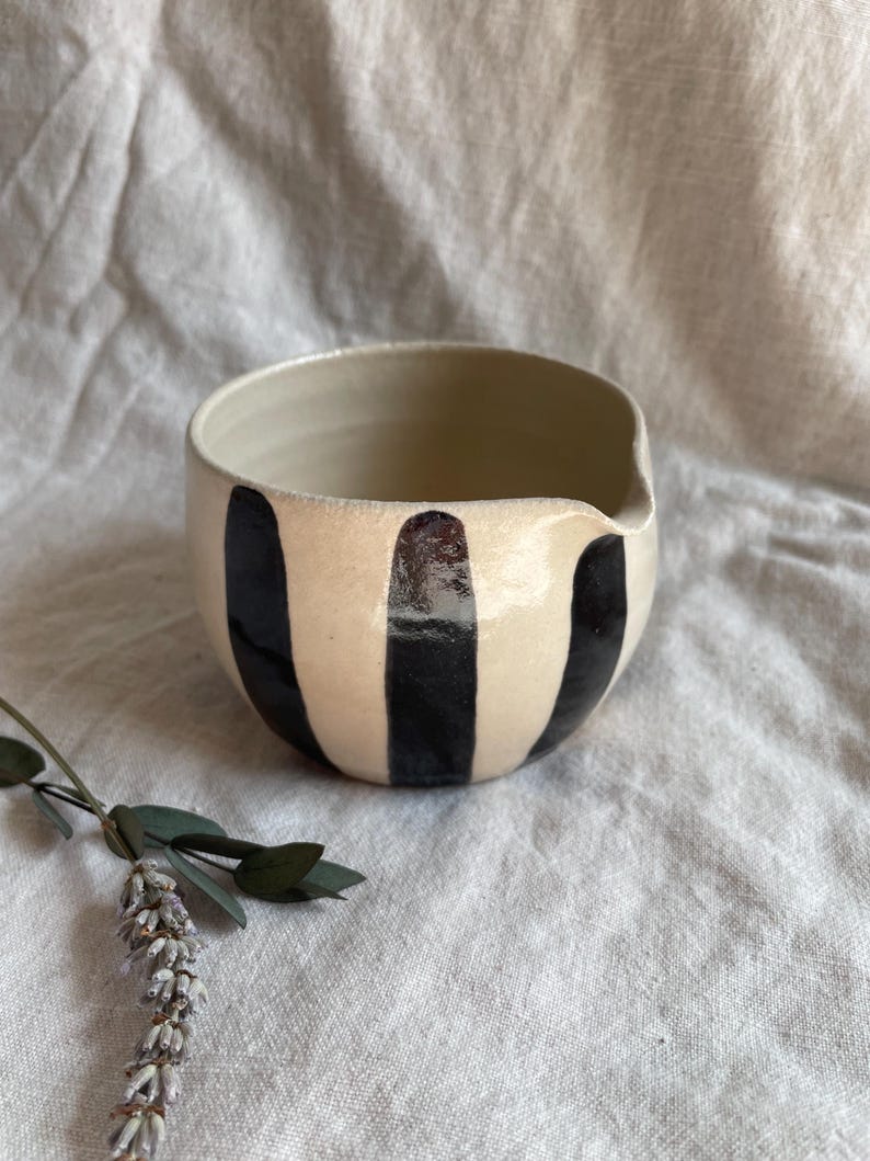 May include: A white ceramic bowl with black vertical stripes. The bowl has a slightly rounded shape and a wide rim.