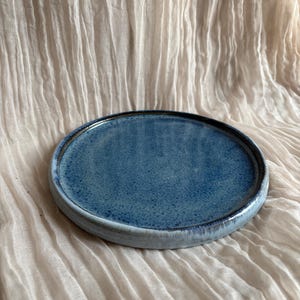 May include: A round, ceramic plate with a speckled blue glaze. The plate has a slightly raised edge and a dark brown rim. The plate is approximately 20 cm in diameter.
