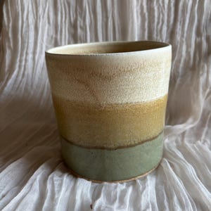 May include: A handcrafted ceramic plant pot with a cylindrical form. The pot displays a colour gradient, beginning with a light beige at the top, shifting to a golden yellow, and finishing with a muted green at the base. Suitable for indoor plants, it is a decorative item.