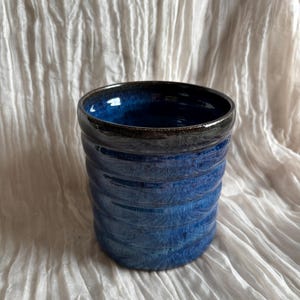 May include: A blue ceramic tumbler with a textured, ridged exterior. The interior is dark blue, and the rim is black. The background is a white, textured fabric. A handcrafted piece.