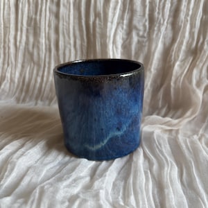 May include: A handcrafted ceramic tumbler with a glossy, deep blue glaze. The cup has a slightly tapered shape and a dark rim. The blue colour transitions from dark at the top to lighter shades, creating a unique, artistic look. The tumbler is set against a white, textured background.