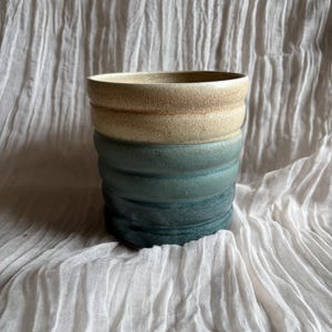 May include: A handmade ceramic vessel with a cylindrical shape. The top section is a light tan color, transitioning to a teal blue at the base. The surface has a textured, ribbed design. The vessel sits on a white, textured fabric.