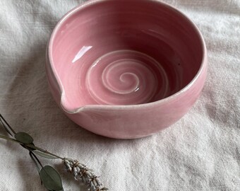 May include: A pink ceramic bowl with a swirl design. The bowl has a small spout on the side.