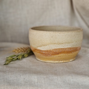 May include: A handmade ceramic bowl with a white glaze and brown and yellow stripes. The bowl is sitting on a beige fabric surface.