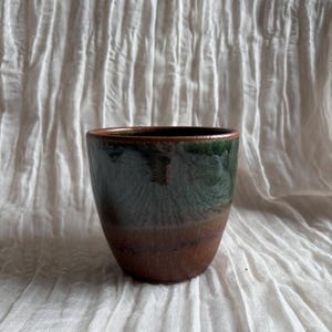 May include: A handmade ceramic cup with a dark green and brown glaze. The cup has a rounded shape and a smooth, glossy finish. The interior is dark, and the rim is a darker shade of green.
