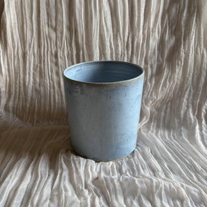 May include: A pale blue ceramic plant pot with a cylindrical form. The pot has a subtly textured finish and a light brown rim. The background is a white, textured fabric. Suitable for indoor or outdoor use, it measures approximately 15 cm in height.