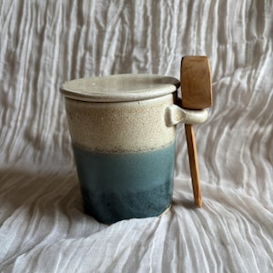 May include: A ceramic container with a lid and a wooden spoon. The container is cylindrical, with a gradient of colours from beige to teal. The spoon rests on a small holder attached to the side.