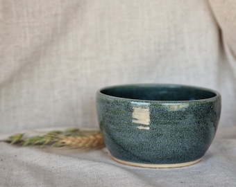 May include: A dark green ceramic bowl with a speckled glaze. The bowl is sitting on a light beige fabric surface.