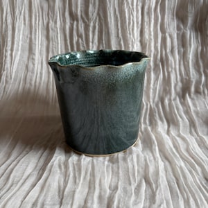 May include: A dark teal ceramic pot with a wavy rim. The pot has a textured surface and a light-coloured base. The interior is a darker shade of teal. The pot is likely used for plants or as a decorative item.