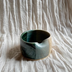 May include: A handcrafted ceramic bowl with a distinctive design. The bowl features teal, green, and tan hues. It has a small spout on one side. The bowl is placed on a white, textured surface.