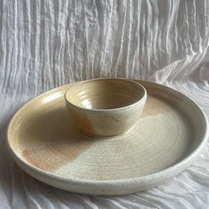 May include: A handcrafted ceramic serving dish set. The set comprises a large, circular platter with a raised edge and a small, circular bowl in the centre. The dish and bowl are a creamy beige colour with a gradient of light brown. Perfect for serving snacks or appetizers.