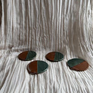 May include: Four ceramic coasters with a two-tone design. Each coaster is split in half, with one side in a deep green and the other in a warm brown. The coasters are round and have a glossy finish. They are arranged on a white, textured fabric.
