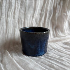 May include: A small, handmade ceramic cup with a dark blue and black glaze. The cup has a cylindrical shape and a smooth, glossy finish. The interior of the cup is a deep blue, and the exterior has a marbled effect. The cup is approximately 7.6 cm tall.