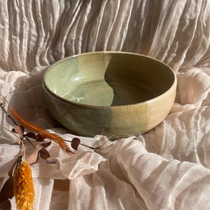 May include: A large, green and brown ceramic bowl with a slightly textured surface. The bowl is sitting on a light brown fabric background.