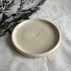 Puede incluir: Un plato peque&ntilde;o, redondo, de cer&aacute;mica de color blanco roto con un borde ligeramente elevado. El plato tiene una superficie sutilmente texturizada.