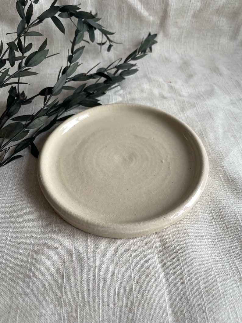Puede incluir: Un plato peque&ntilde;o, redondo, de cer&aacute;mica de color blanco roto con un borde ligeramente elevado. El plato tiene una superficie sutilmente texturizada.