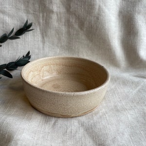 May include: A small, off-white ceramic bowl with a speckled glaze. The bowl is round and has a slightly flared rim.