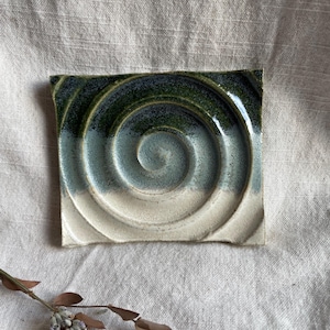 May include: A square ceramic tile with a white, blue, and green glaze. The tile has a spiral design that is raised and textured.