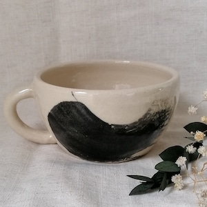 May include: A white ceramic mug with a black abstract design. The mug has a rounded shape and a handle.
