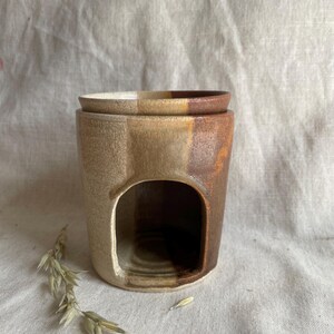 May include: A ceramic oil warmer in beige, brown, and orange. The cylindrical warmer has an arched opening and a matching lid. A sprig of dried grass lies beside the warmer. The warmer is likely used for aromatherapy.
