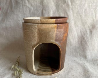 May include: A ceramic oil warmer with a two-tone design in shades of beige, brown, and orange. The warmer has a cylindrical base with an arched opening and a matching lid. A sprig of dried grass rests beside the warmer.