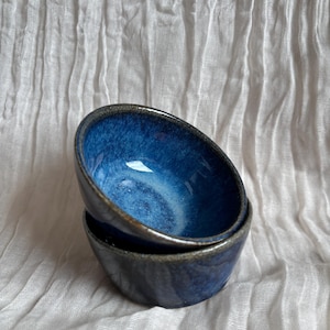 May include: Two small, handmade ceramic bowls stacked. The bowls have a deep blue interior with a glossy finish and a dark rim. The exterior is a darker blue. The bowls are set against a white, textured background.
