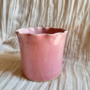 May include: A pink ceramic vase with a ruffled rim. The vase has a cylindrical shape and a smooth, glossy finish. The color is a soft, muted pink. The vase is likely handmade and suitable for holding flowers or as a decorative item.