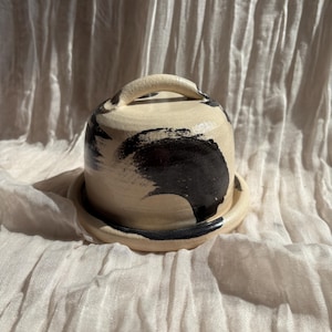 May include: A ceramic butter dish with a lid and handle. The dish is off-white with black brushstroke designs. The lid has a rounded shape and a small handle. The dish sits on a matching saucer.
