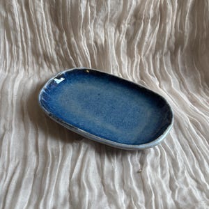 May include: A handmade, oval ceramic dish with a glossy, deep blue glaze. The dish has an irregular shape and a light blue rim. The background is a textured, off-white fabric.