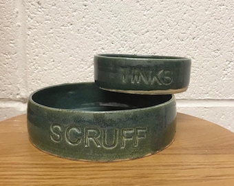 May include: Two green ceramic dog bowls, one stacked on top of the other. The larger bowl says "Scruff" and the smaller bowl says "Tinks".