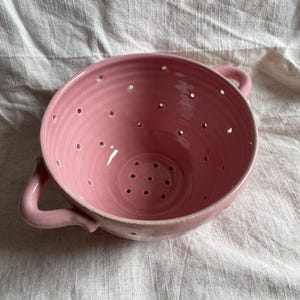 May include: A round, light pink ceramic colander featuring two curved side handles and a series of drainage holes distributed across the bowl and base. The interior and exterior surfaces have a smooth, glossy finish with visible pottery wheel rings.