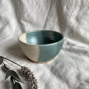 May include: A small, white and blue ceramic bowl with a speckled glaze. The bowl is sitting on a white fabric surface.