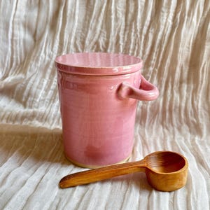 Könnte beinhalten: Ein rosa Keramikbehälter mit Deckel und zwei kleinen Griffen, zusammen mit einem Holzschöpfer. Der Behälter ist zylindrisch und hat eine glänzende Oberfläche. Der Holzschöpfer ist hellbraun und hat eine abgerundete Schale. Die Artikel befinden sich auf einer strukturierten, cremefarbenen Oberfläche.