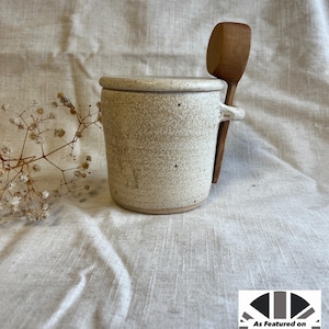 May include: White ceramic jar with a speckled glaze and a wooden spoon resting on the rim. The jar has a lid and a handle. The jar is sitting on a white fabric surface.