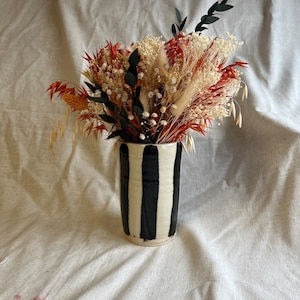 May include: A cylindrical ceramic vase with vertical black stripes on a cream-coloured base, filled with a dried floral arrangement featuring red, white, and black stems, including grasses, baby's breath, and dark eucalyptus leaves.
