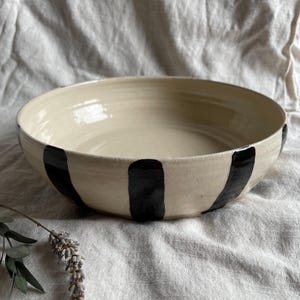 May include: A large, white ceramic bowl with black stripes. The bowl is sitting on a white fabric surface.