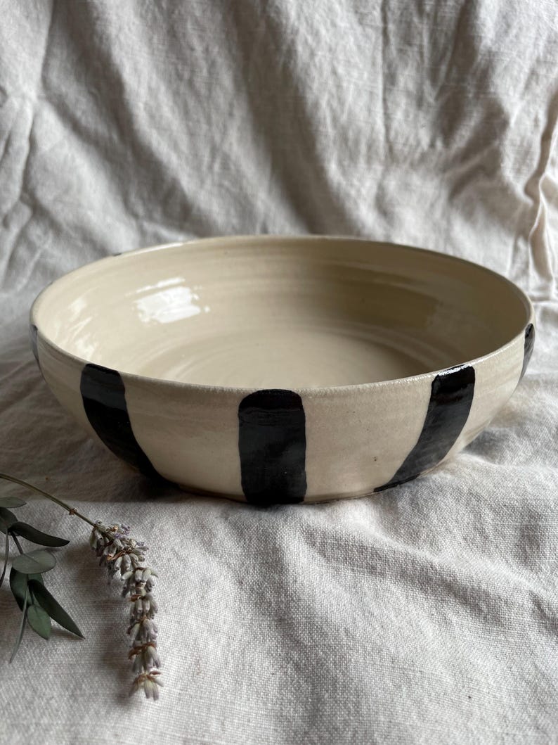 May include: A large, white ceramic bowl with black stripes. The bowl is sitting on a white fabric surface.