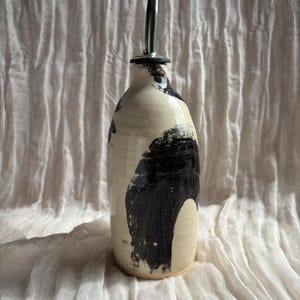 May include: A ceramic oil bottle with a stainless steel spout. The bottle is off-white with large, abstract black brushstrokes. The design is minimalist and modern, suitable for kitchen decor.