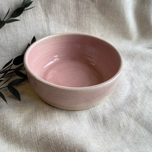 May include: A light pink ceramic bowl with a slightly textured surface. The bowl is empty and sitting on a light beige fabric background.