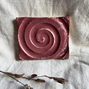 May include: A square, handmade ceramic soap dish in a deep burgundy color. The dish features a raised spiral design in the center and is set against a neutral linen background. The edges have a slightly textured, natural finish.