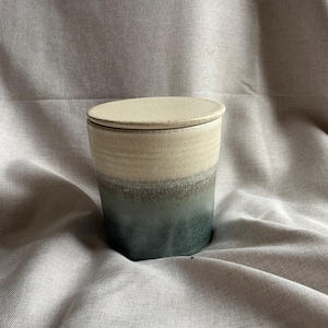 May include: A ceramic jar with a lid. The jar is cylindrical with a gradient of colors, transitioning from beige at the top to a teal-green at the base. The lid is a matching beige color. The jar is sitting on a neutral-colored fabric.