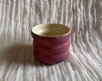 May include: A handmade, small ceramic cup with a textured, ribbed design. The cup is a deep magenta color with a cream-colored interior. The rim has a subtle, dark purple accent. The cup is likely used for drinking beverages.