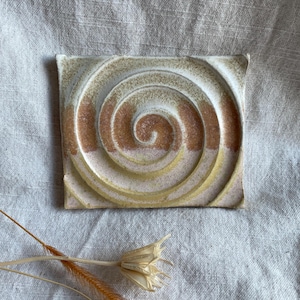 May include: A square ceramic coaster featuring a raised spiral design in shades of white, beige, and brown.