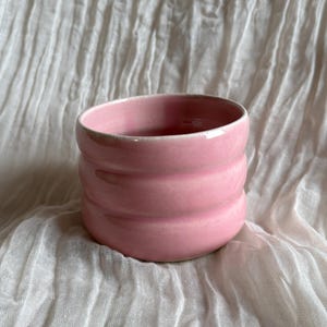 May include: A pink ceramic cup with a ribbed design. The cup is a soft, pastel pink colour and has a smooth, glossy finish. The cup is sitting on a white, textured surface.